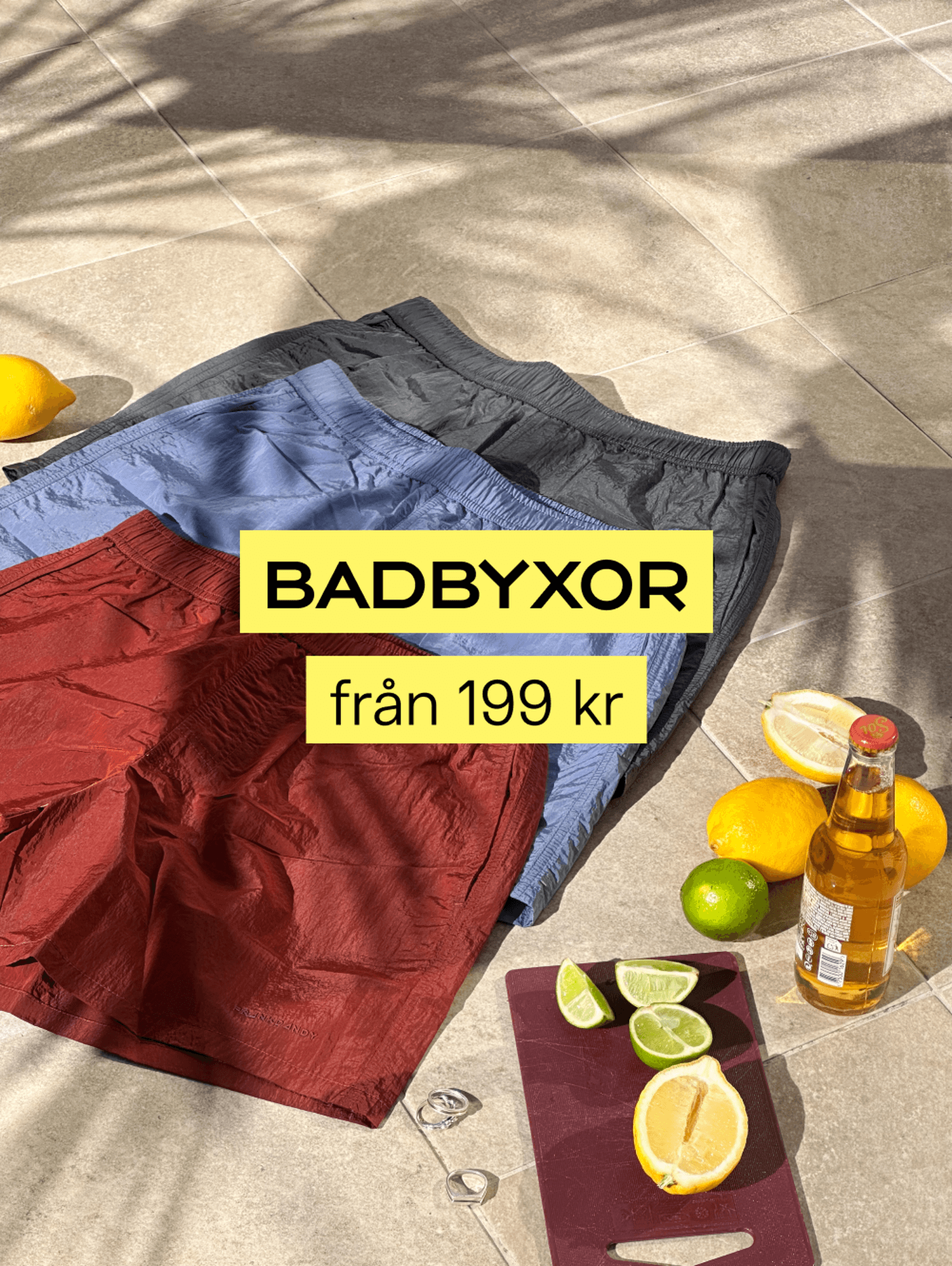 Three pairs of swim trunks on a tiled floor with lemons, lime, a beer bottle, and a cutting board. Text: "BADBYXOR från 199 kr".