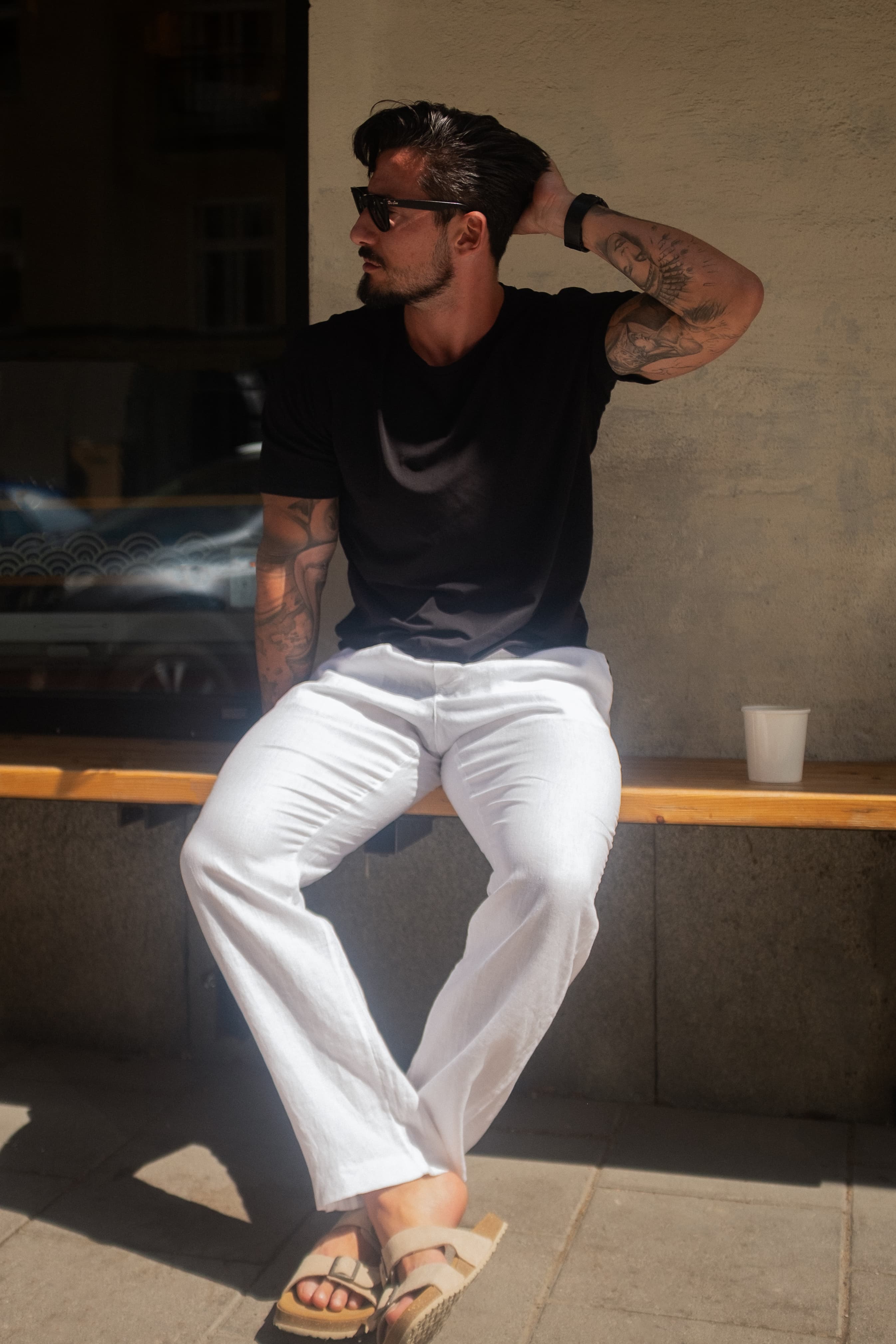 Man in sunglasses and tattoos sits on a bench, wearing a black shirt and white pants, with one hand behind his head, in sunlight.